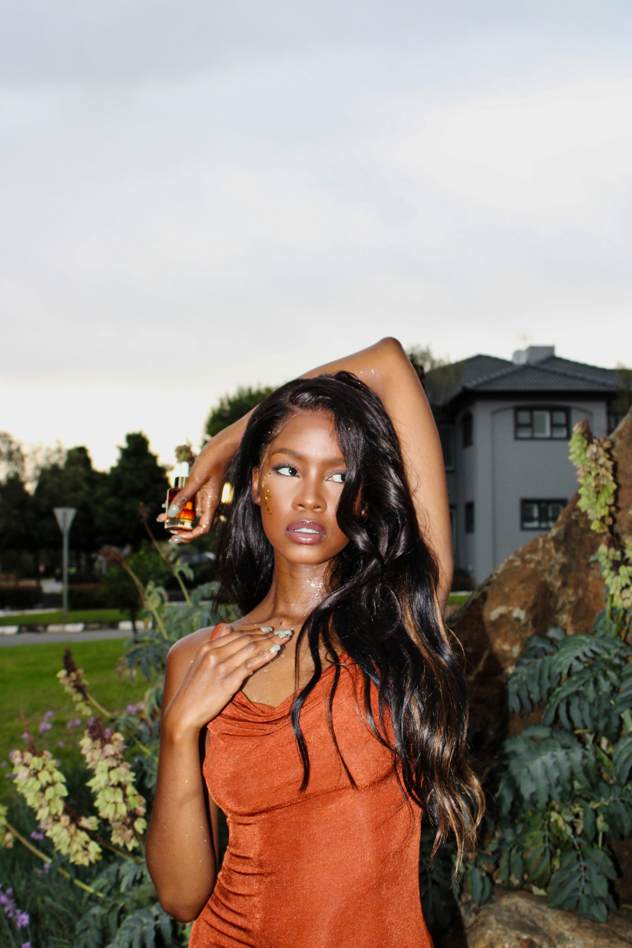 Model in a copper satin dress holding a Luxe Elixir Cosmetics perfume oil bottle above her head at dusk