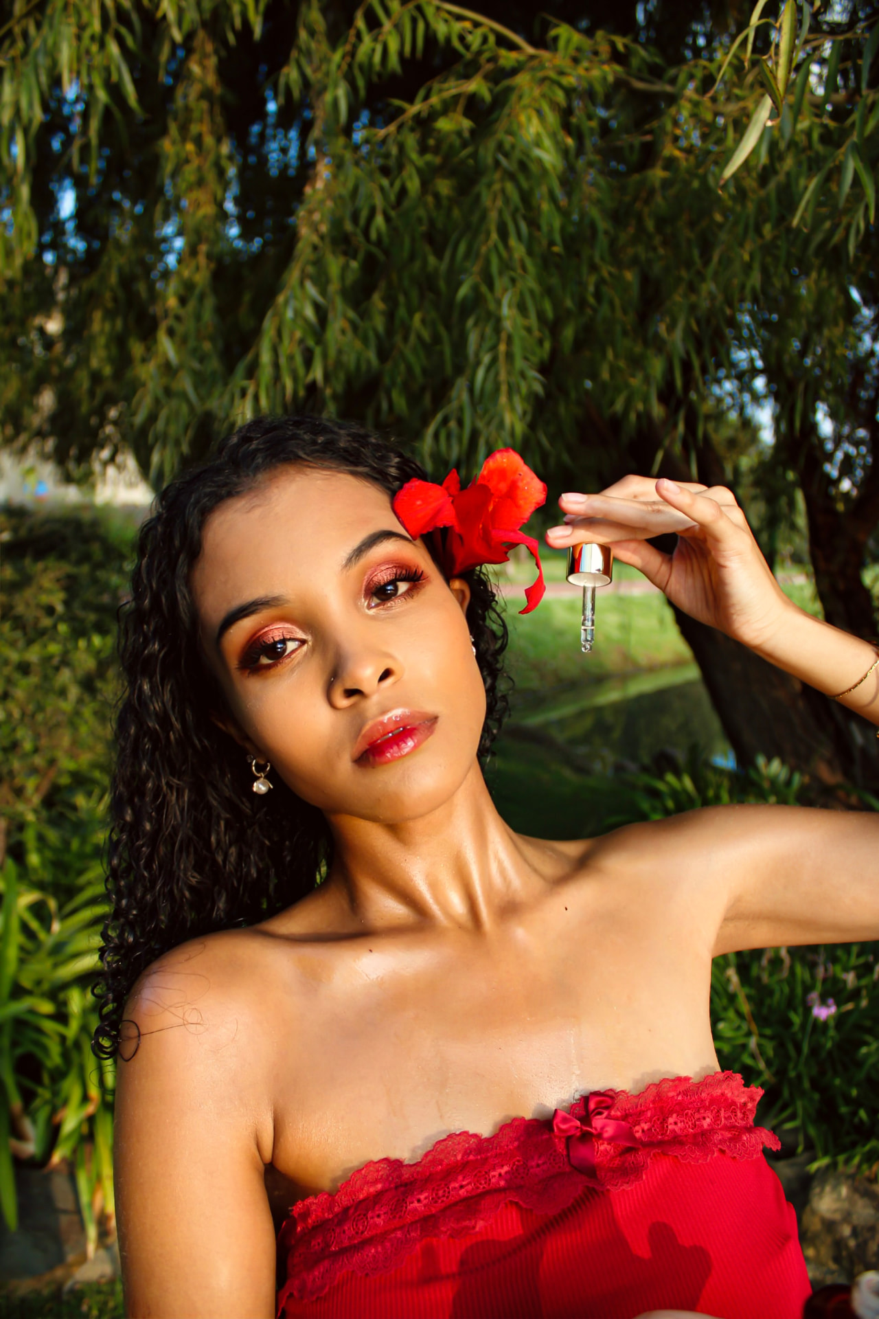 Model with a red hibiscus in her hair holding a perfume oil dropper beneath a willow tree