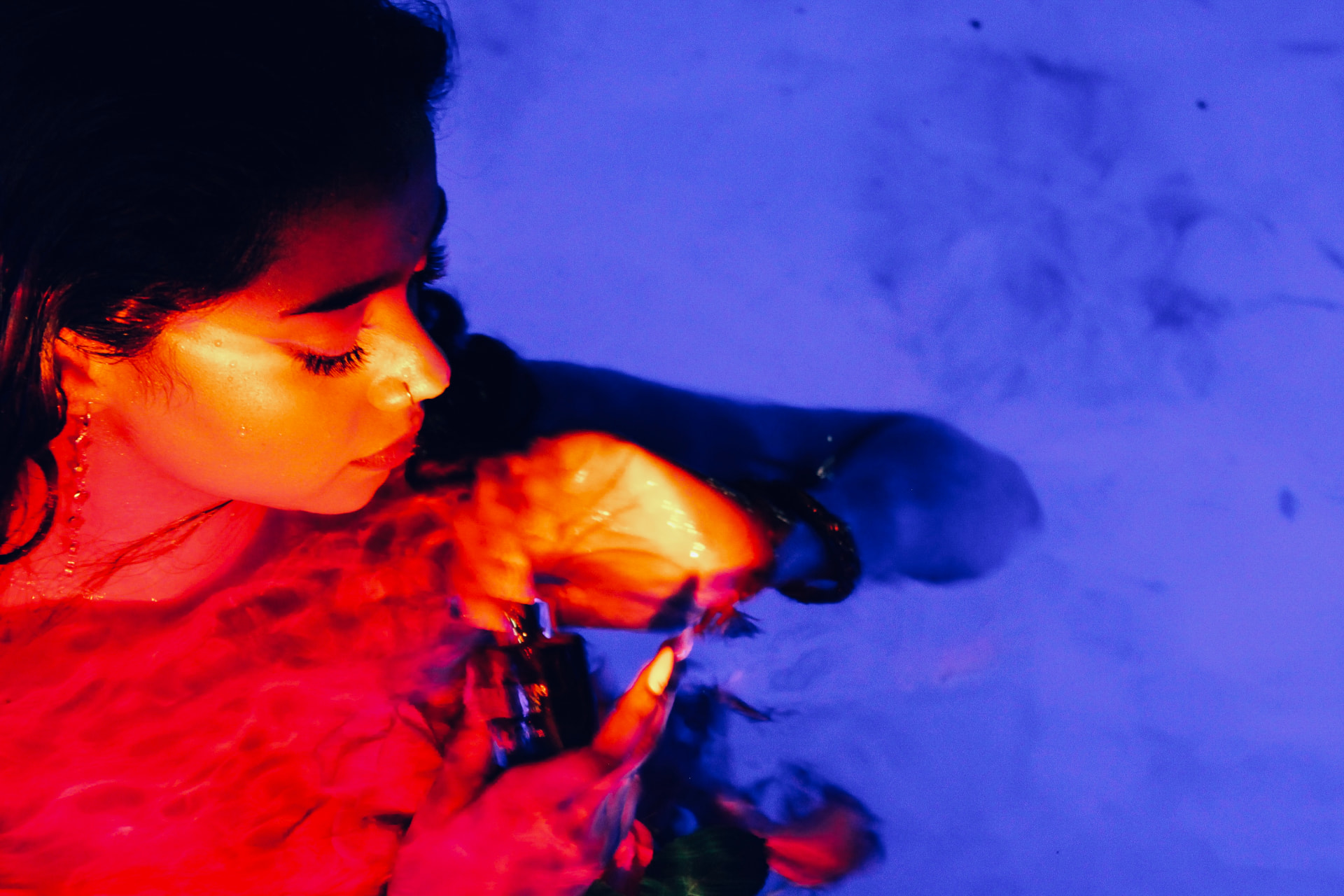 Editorial portrait of a model in water lit in red and blue, cradling a perfume oil bottle